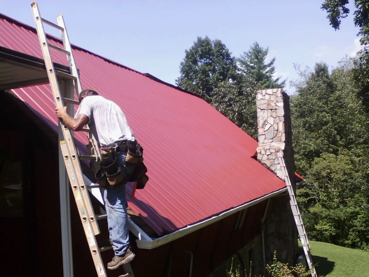Roofer on a ladder beside a red metal roof and stone chimney — Soffit & Fascia Repair by Roofing Brothers in Henryetta
